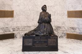 A wide shot of the Mother Joseph statue, which is made of bronze and depicts a nun kneeling in prayer. The statue sits on a granite base in the Legislative Building foyer.