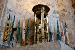 A bronze firepot with Roman-style designs. Light shines from the top of the firepot, which stands in front of 10 Washington state county flags.