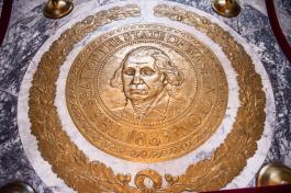A giant, polished bronze Washington State Seal framed by a polished bronze oak leaf wreath placed on a marble floor. Red ropes surround the bronze seal, protecting it from foot traffic.