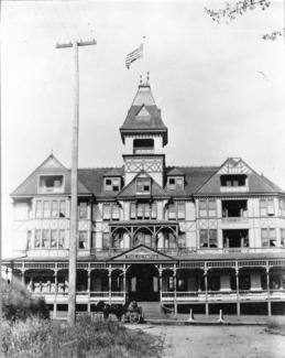 A black and white image of the front of the old Olympia Hotel, which is four stories tall with a tall tower in the center. An American Flag waves at the top of the tower, and a horse carriage is parked in front of the hotel.