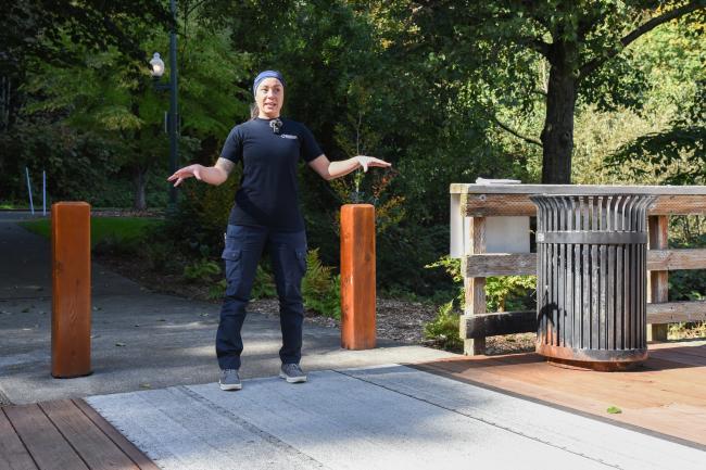 A woman talking about and gesturing toward the deck at the Capitol Lake Interpretive Center. She stands between two wooden bollards, with large green trees and a garbage can in the shot.