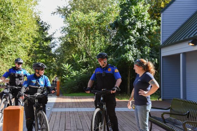 Three Washington State Patrol officers on bikes talking to a woman near the entrance of the Capitol Lake Interpretive Center. Large green trees are behind the group, and a restroom building is to their right.