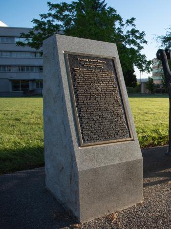 The George Bush Monument, a bronze plaque set in a granite slab, next to a metal park bench with two large trees and the General Administration building in the background.