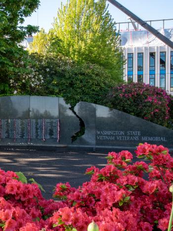 A patch of beautiful red flowers in the foreground surrounds the granite Vietnam Veterans Memorial, featuring small American flags hanging next to the names of Washington state residents killed or missing during the Vietnam War, which is shaded by the trees behind it.