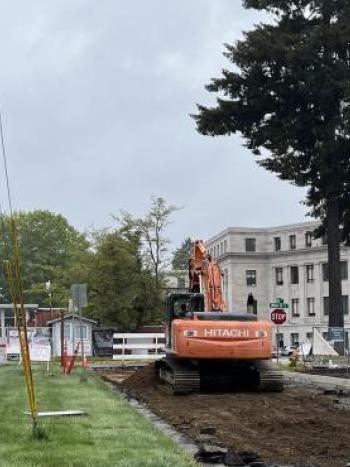 Heavy equipment being driven on an unpaved road with the Legislative Building in the background.
