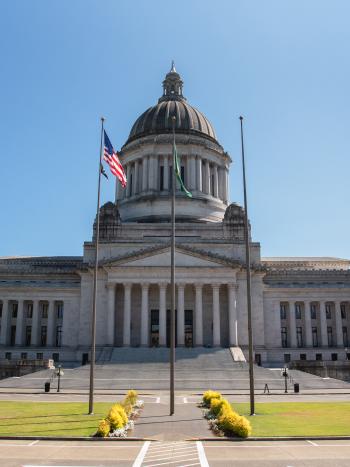 A wide shot of the Legislative Building on a sunny day, with flag poles in front of it and large green trees surrounding it.