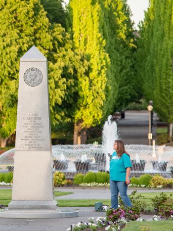 A man and a woman standing on both sides of the Medal of Honor Memorial, reading its inscriptions, with the Tivoli Fountain and green trees in the background.