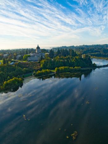 An aerial view of a blue Capitol Lake surrounded by green trees, with the Legislative Building and parts of Capitol Campus in the distance.