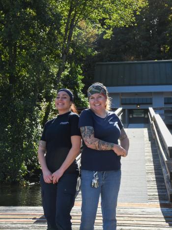 Two women, Angela Gongora-Hines and Kailee Moulton, standing on a wooden deck at the Capitol Lake Interpretive Center, surrounded by large green trees.