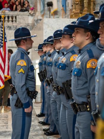 A police officer in a blue uniform facing a row of other police officers, with the Washington State and American Flag on each side of the row. The marble interior of the Legislative Building rotunda and a government official in a suit are in the background.