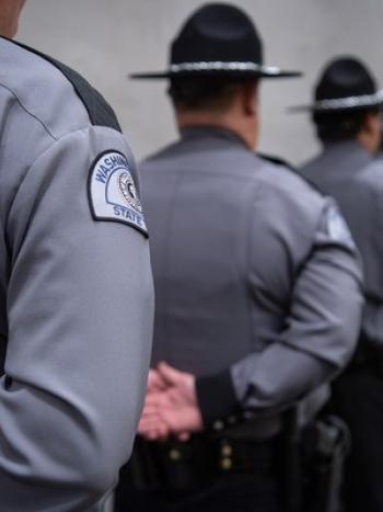 A close up of a Washington State Patrol patch on an officer's uniform. The officer is in a line with other police officers wearing gray shirts and black hats, all standing in the "at ease" stance.