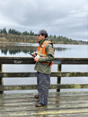 A man with a reflective orange vest is standing on a wooden deck over Capitol Lake while collecting data in a smartphone and a notepad.