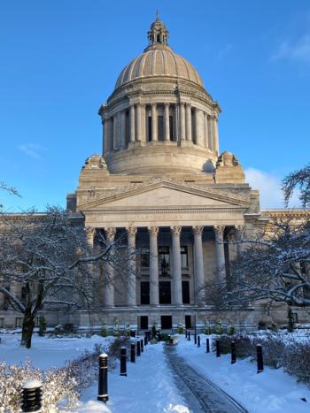 A snow-covered walkway and lawn facing the south side of the Legislative Building. A grounds crew member operates a small snowplow in the distance.