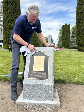 DeWayne Steinman, a DES Buildings & Grounds employee, is shown polishing the new bronze plaque that rests atop a granite base on the Washington State Capitol Campus with trees, bushes, grass, and blue sky in the background.