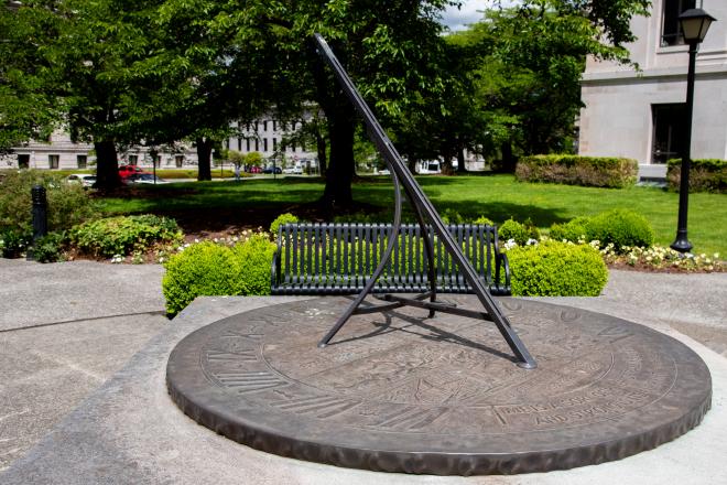 An image of the Territorial Sundial with a bench and trees in the background.