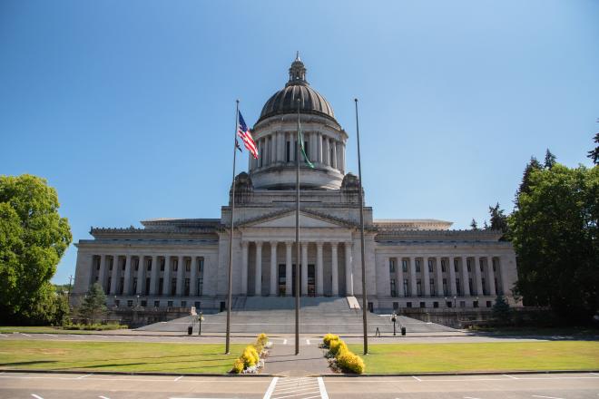 A wide shot of the Legislative Building with parked cars and flag poles in front of it and trees surrounding it.