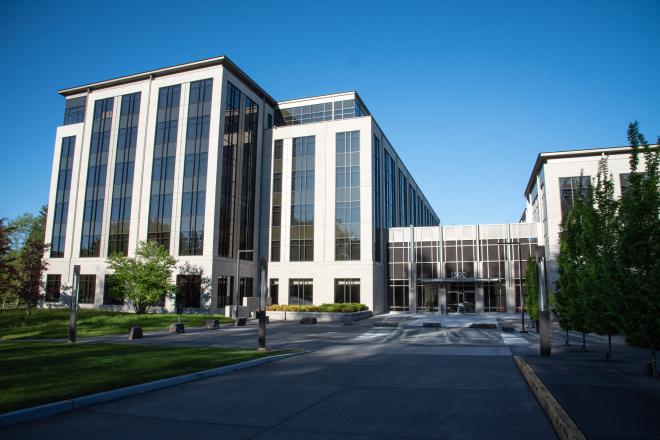 A wide shot of the 1500 Jefferson building, which is a six-floor office building with a predominantly white exterior and large, dark windows. There are small green trees and green grass surrounding the driveway leading to the building's entrance.