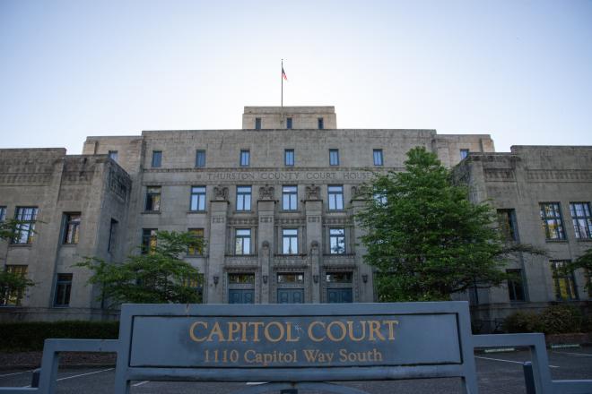 A wide shot of the Capitol Court Building, which is a four-story stone building with large blue doors and an American flag waving on the roof. The words "Thurston County Court House" are inscribed in the stone near the top of the building, and there is a blue sign with gold lettering in the foreground that says, "Capitol Court, 1110 Capitol Way South."