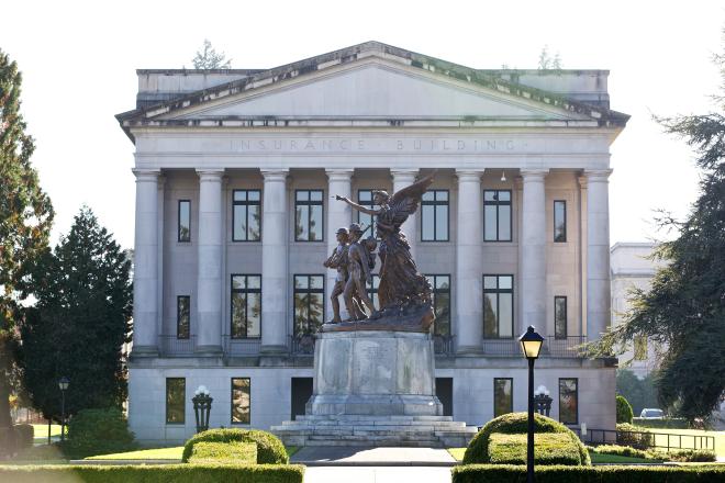 The front of the Insurance Building, which was built with white stone in a classical architecture style. Eight cylinder-shaped pillars frame the entrance to the building, and the bronze Winged Victory Monument stands tall in the roundabout in front of the building.