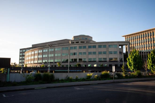 A wide shot of the Natural Resources Building, one of the largest buildings on Capitol Campus, which is predominantly made of concrete. The Natural Resources Building parking garage is in the foreground and the Highways-Licenses Building is in the background, with various trees and other plants scattered between the areas.