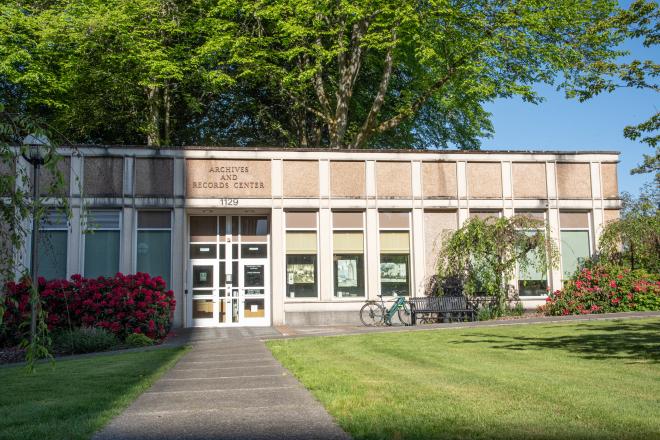 The entrance of the State Archives building, which is a one-story building made with white and brown stone. A stone pathway lined with flowering bushes, small trees, a bench, and light posts leads up to the building entrance.