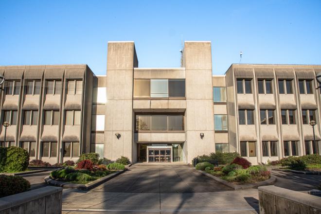 The front of the Department of Transportation Building, which is a three-story concrete building with large rectangular pillars framing the entrance. A concrete path lined with shrubs and flowering bushes leads to the entrance.