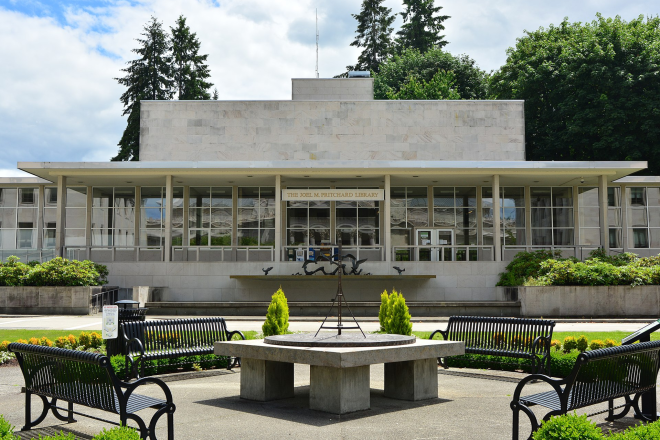 A view of the Joel M. Pritchard Building's front side, which is mainly made of white stone with large glass walls. A fountain, a sundial, four metal park benches, and various green bushes are in front of the building.