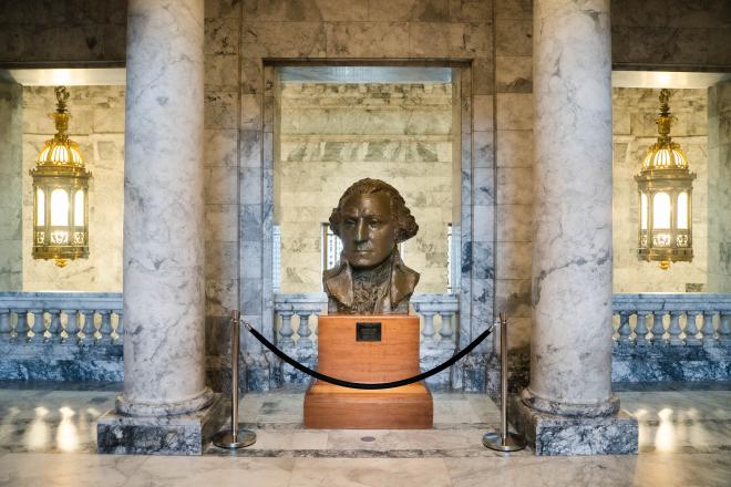 A large bronze statue of President George Washington's head, which sits on a wooden pedestal behind a rope barrier. Two marble columns stand on each side of the statue, and two large bronze chandeliers light up the area.