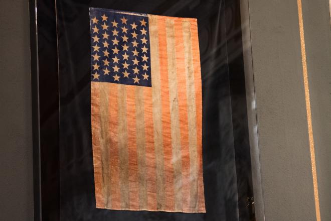 An old, faded American flag with 42 stars hanging on a black platform behind a glass case.