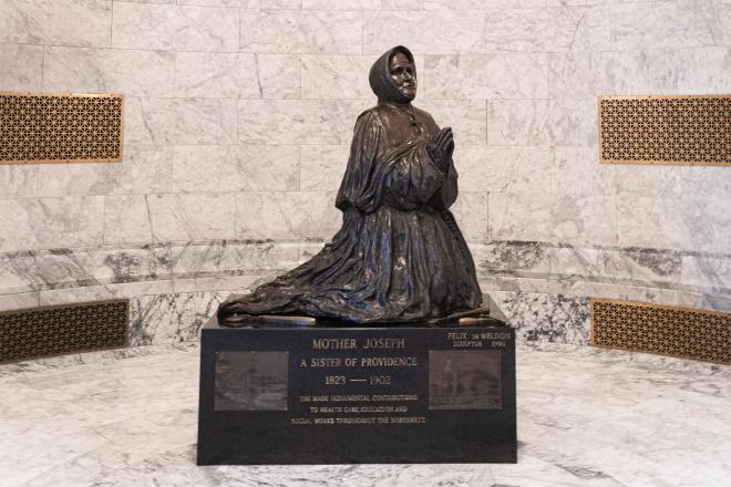 A wide shot of the Mother Joseph statue, which is made of bronze and depicts a nun kneeling in prayer. The statue sits on a granite base in the Legislative Building foyer.