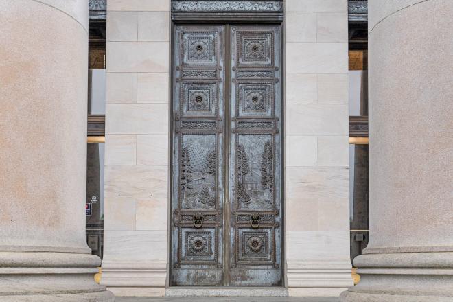 A set of bronze doors at the front of the Legislative Building. The dark bronze has some blueish green spots due to weathering but remains in overall good condition.
