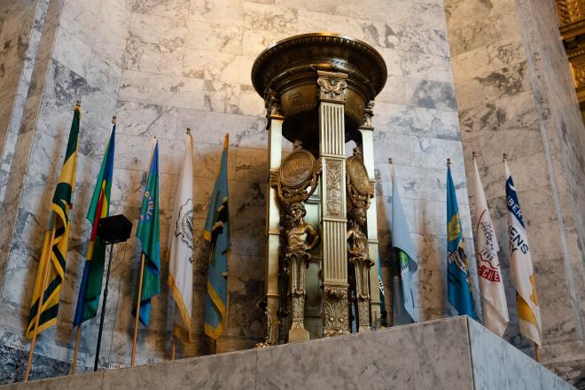 A bronze firepot with Roman-style designs. Light shines from the top of the firepot, which stands in front of 10 Washington state county flags.