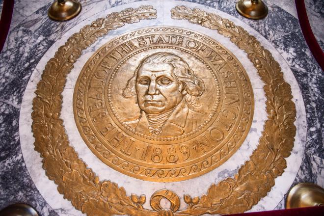 A giant, polished bronze Washington State Seal framed by a polished bronze oak leaf wreath placed on a marble floor. Red ropes surround the bronze seal, protecting it from foot traffic.